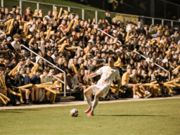 UMBC Men's soccer player kicking a ball at a night game with UMBC student fans in the background