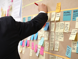 A person's arm holding a pen and writing on a sticky note on a cork board full of sticky notes.