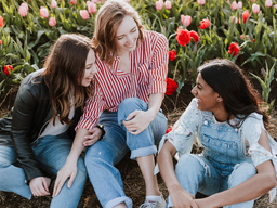 Three young women wearing jeans and shirts sitting on the grass with tulips in the background.