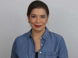 A Latinx woman wearing a blue button up top is smiling. She has on red lipstick.