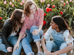 Three young women wearing jeans and shirts sitting on the grass with red and pink tulips in the background.