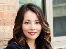 Clarissa Cheah in a blue shirt and chunky colorful necklace in front of a red bricked wall and window
