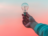 a hand holding a lighbulb with a pink and blue sky behind